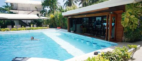 A swimming pool with a person swimming, a bar area with a view of the pool, and a building with a balcony.