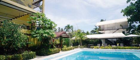 A pool area with a clear blue pool, surrounded by lush greenery and a multi-story building with balconies.