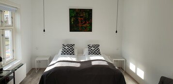 Un dormitorio con cama, dos mesitas de noche, un radiador, una ventana y un cuadro en la pared.