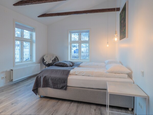 Un dormitorio con una cama, dos ventanas, una silla y una mesita de noche.