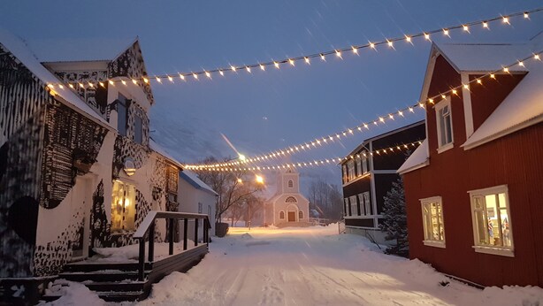 Una calle cubierta de nieve con edificios a ambos lados, luces colgantes arriba y una iglesia a lo lejos.