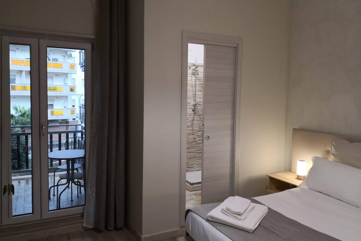 A hotel room with a bed, a bedside table with a lamp, a balcony with a table and chairs, and a view of the building's exterior.