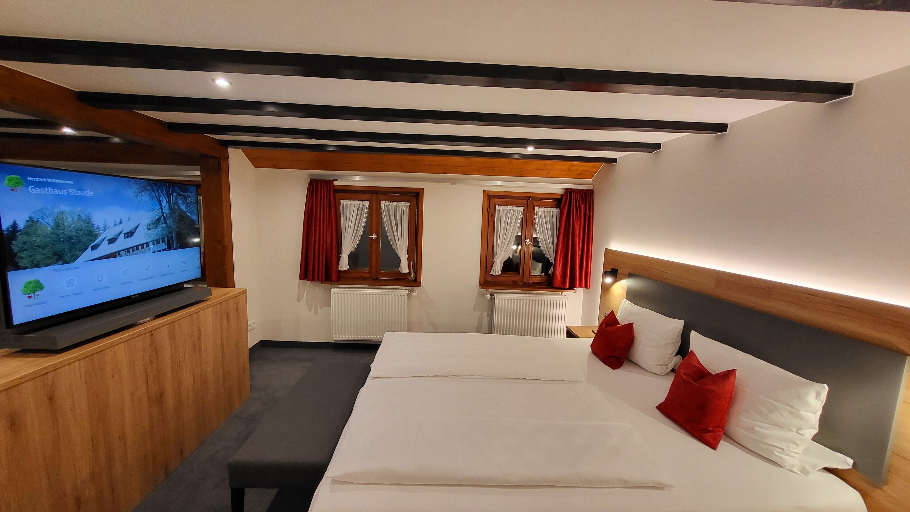 Una habitación de hotel con una cama, un televisor sobre un mueble de madera y dos ventanas con cortinas.