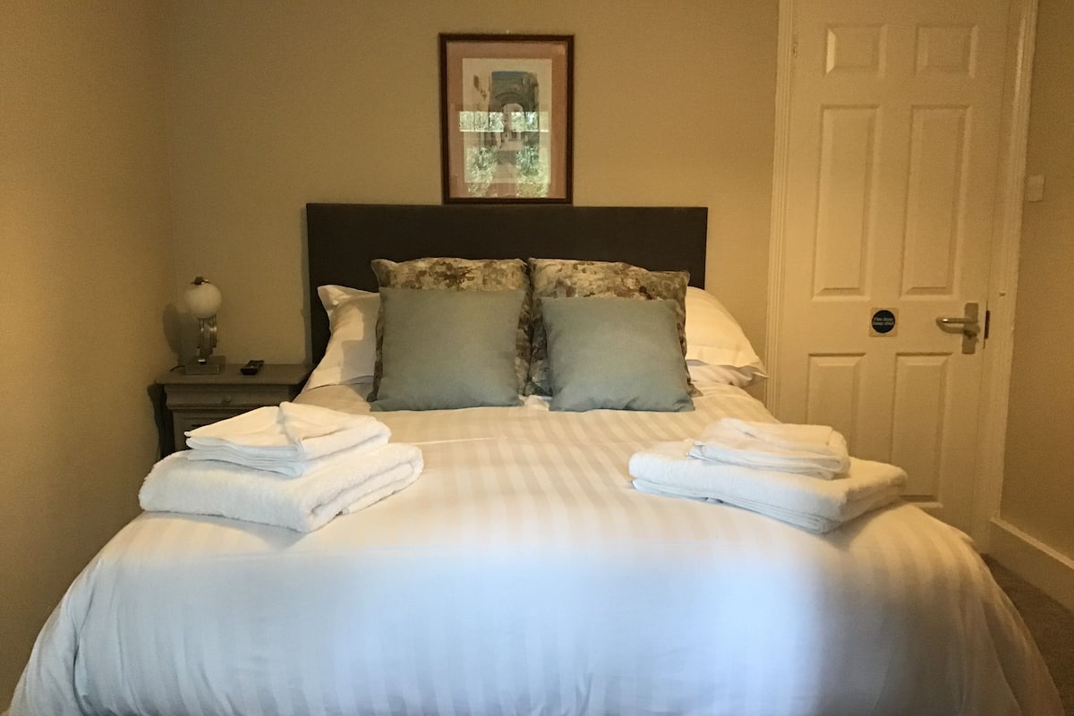 A neatly made bed with pillows and folded towels, a bedside table with a lamp, and a framed picture above the bed.