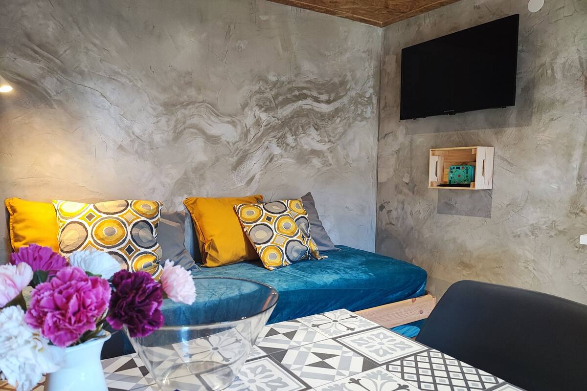 A room with a bed, a TV mounted on the wall, a wooden shelf, and a table with a vase of flowers.