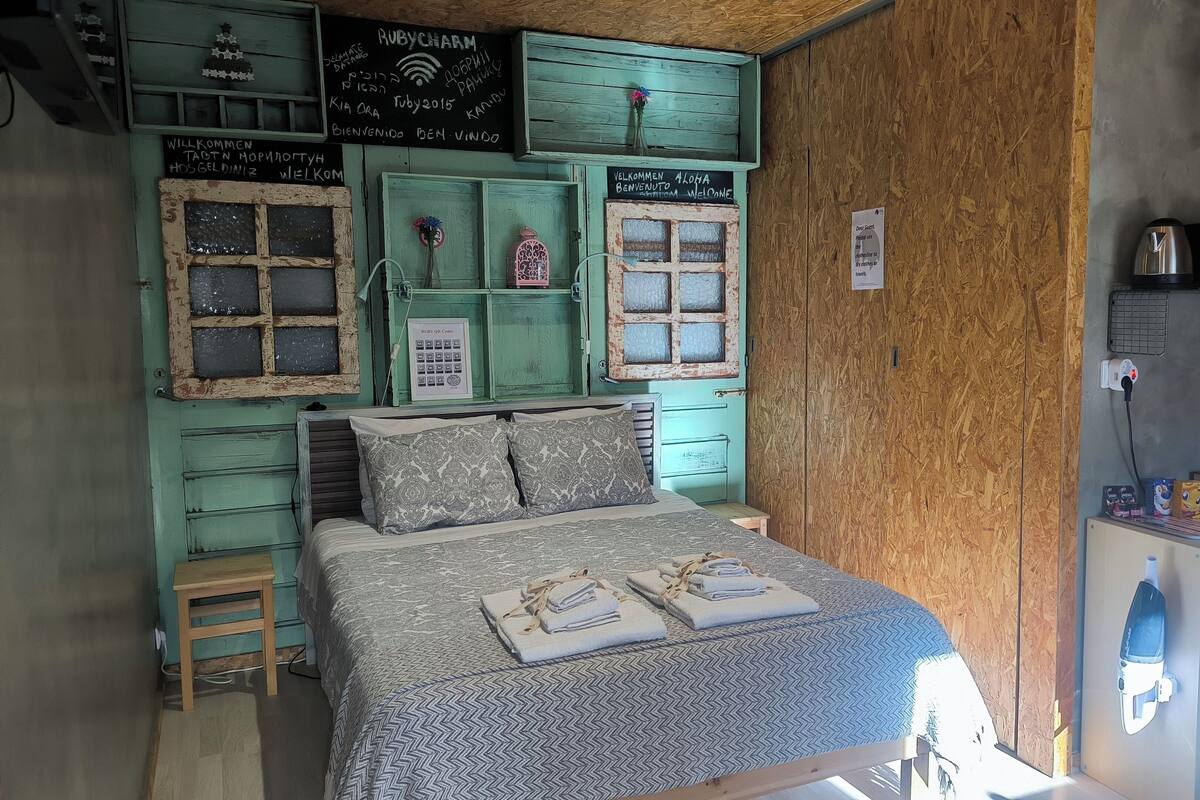 A cozy room with a bed, a small table, and a wooden cabinet. There are signs on the walls in multiple languages, a power outlet, and a kettle on the cabinet.
