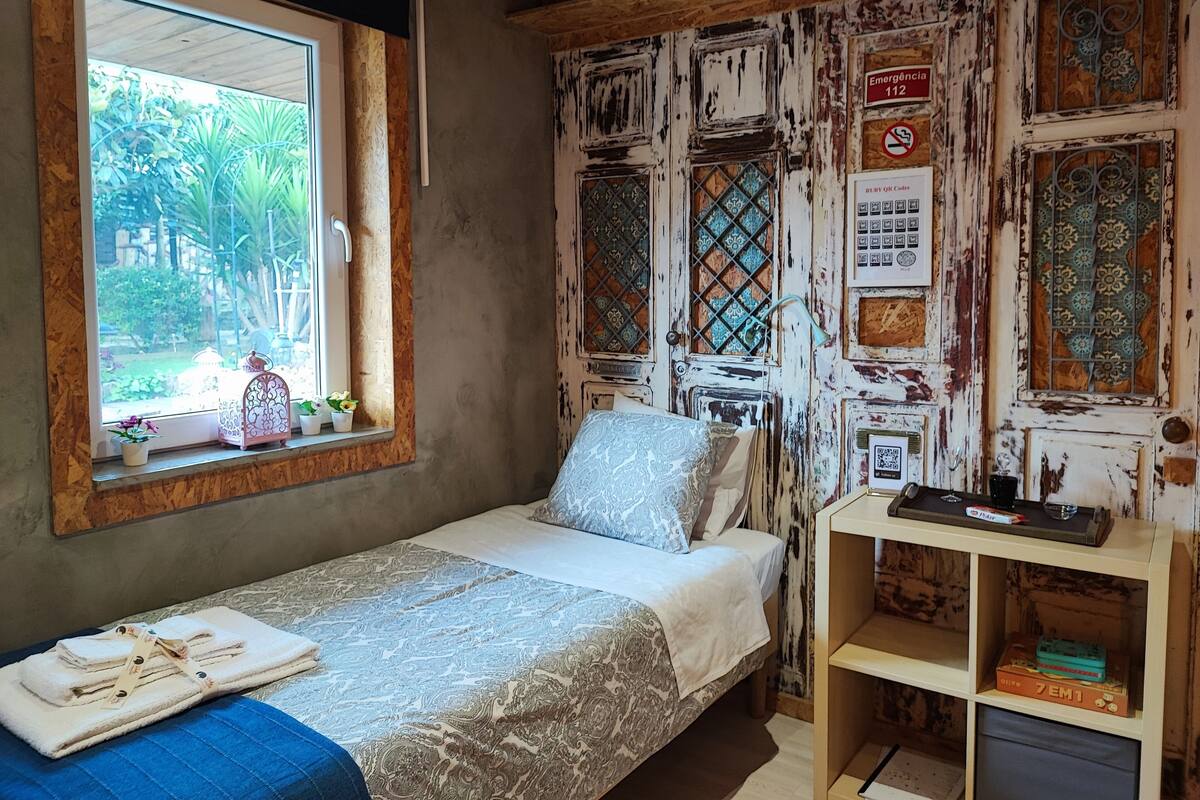 A bedroom with a bed, a window with plants, a wooden shelf, and a wall with various signs and decorations.