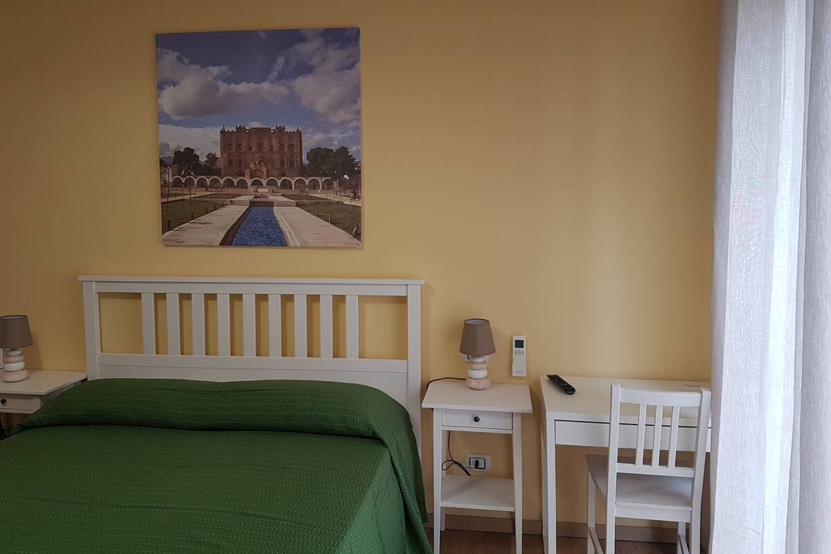 A bedroom with a bed, a desk, a chair, a lamp, and a painting of a castle.