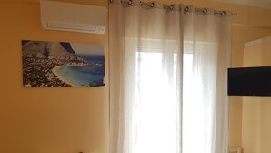 Una habitación con un escritorio blanco, una silla, un ventanal con cortinas translúcidas y un aire acondicionado de pared.