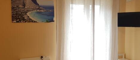 A room with a white desk, a chair, a large window with sheer curtains, and a wall-mounted air conditioner.