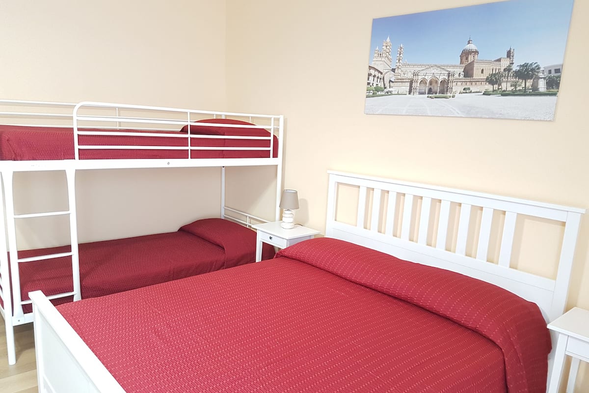 A room with bunk beds, a small bedside table with a lamp, and a painting of a building on the wall.
