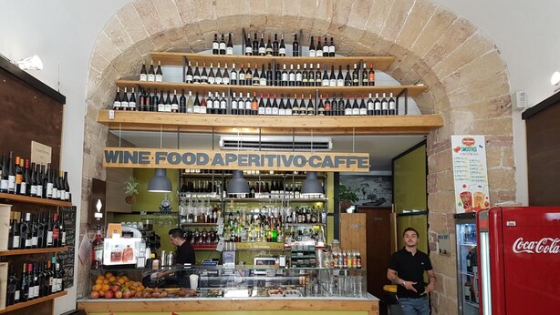 Un bar de vinos con estantes llenos de botellas, una barra con comida y bebidas, y un refrigerador de Coca-Cola.