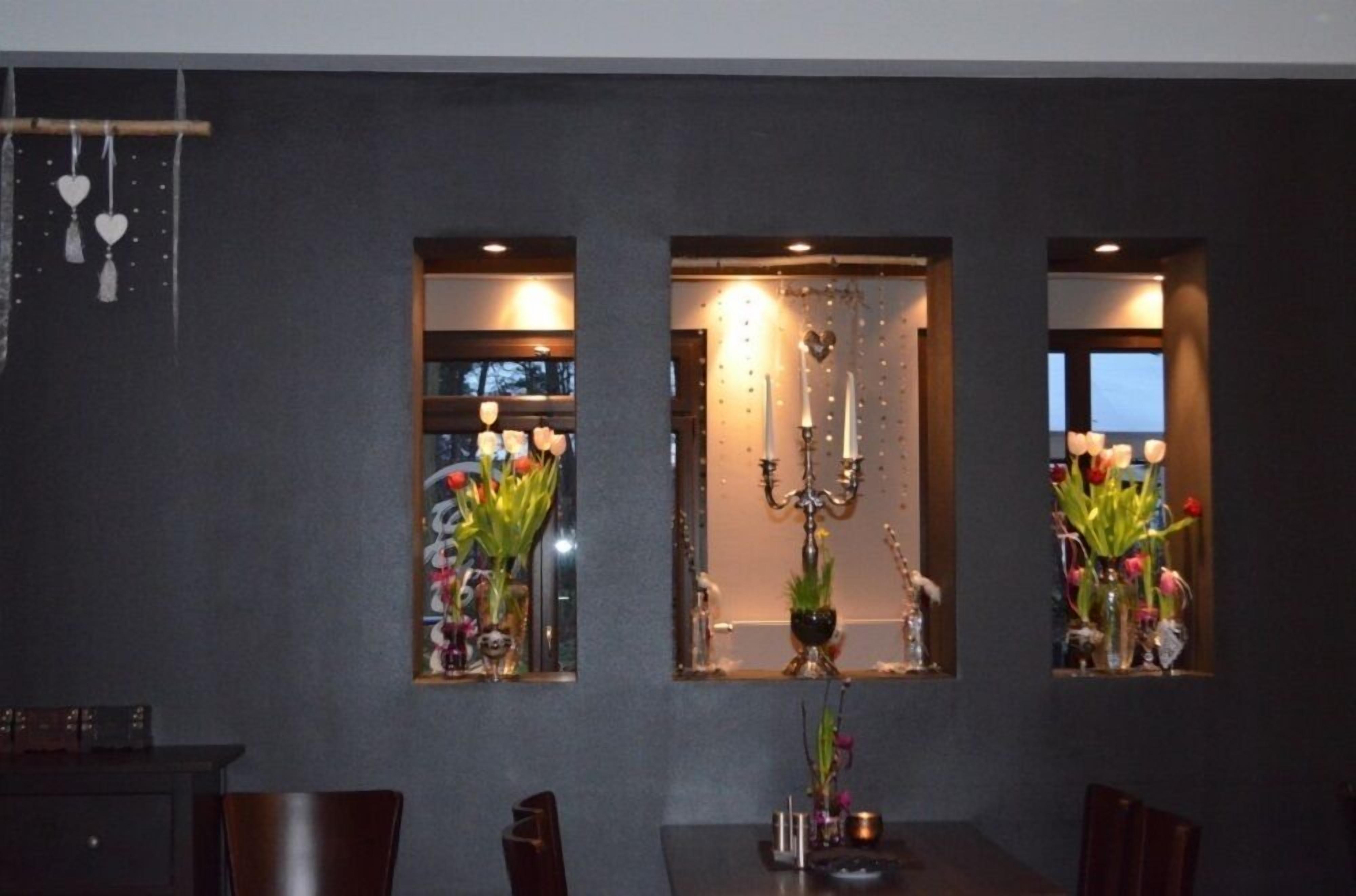 Ein Essbereich mit dunkler Wand, drei Fenstern mit Blumenarrangements und einer Kristallleuchter.