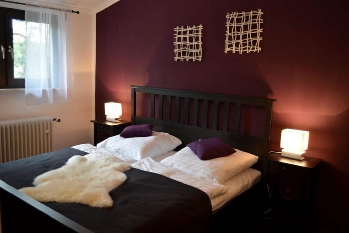 Quarto com cama, duas mesas de cabeceira, janela com cortinas e obras de arte na parede.