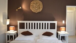 Un dormitorio con una cama blanca, dos mesitas de noche con lámparas, un adorno en la pared y una puerta al fondo.