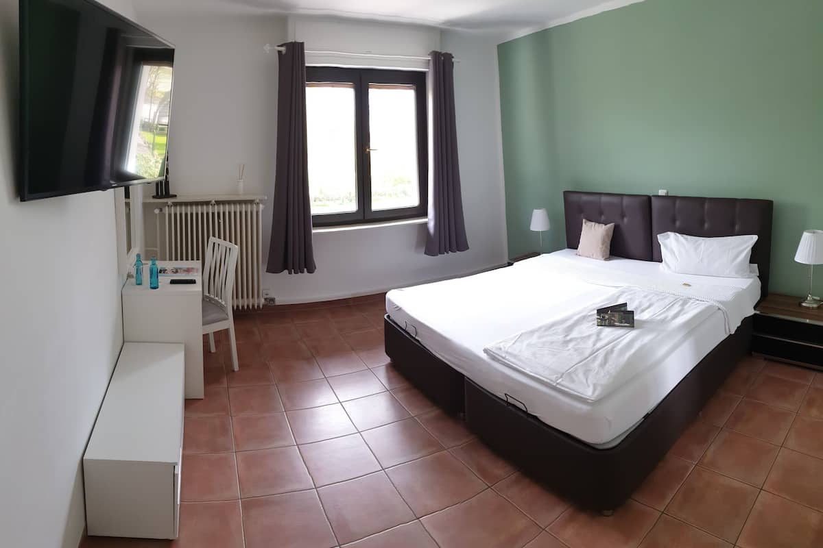 Quarto de hotel com cama, escrivaninha, cadeira, radiador, janela com cortinas e televisão na parede.