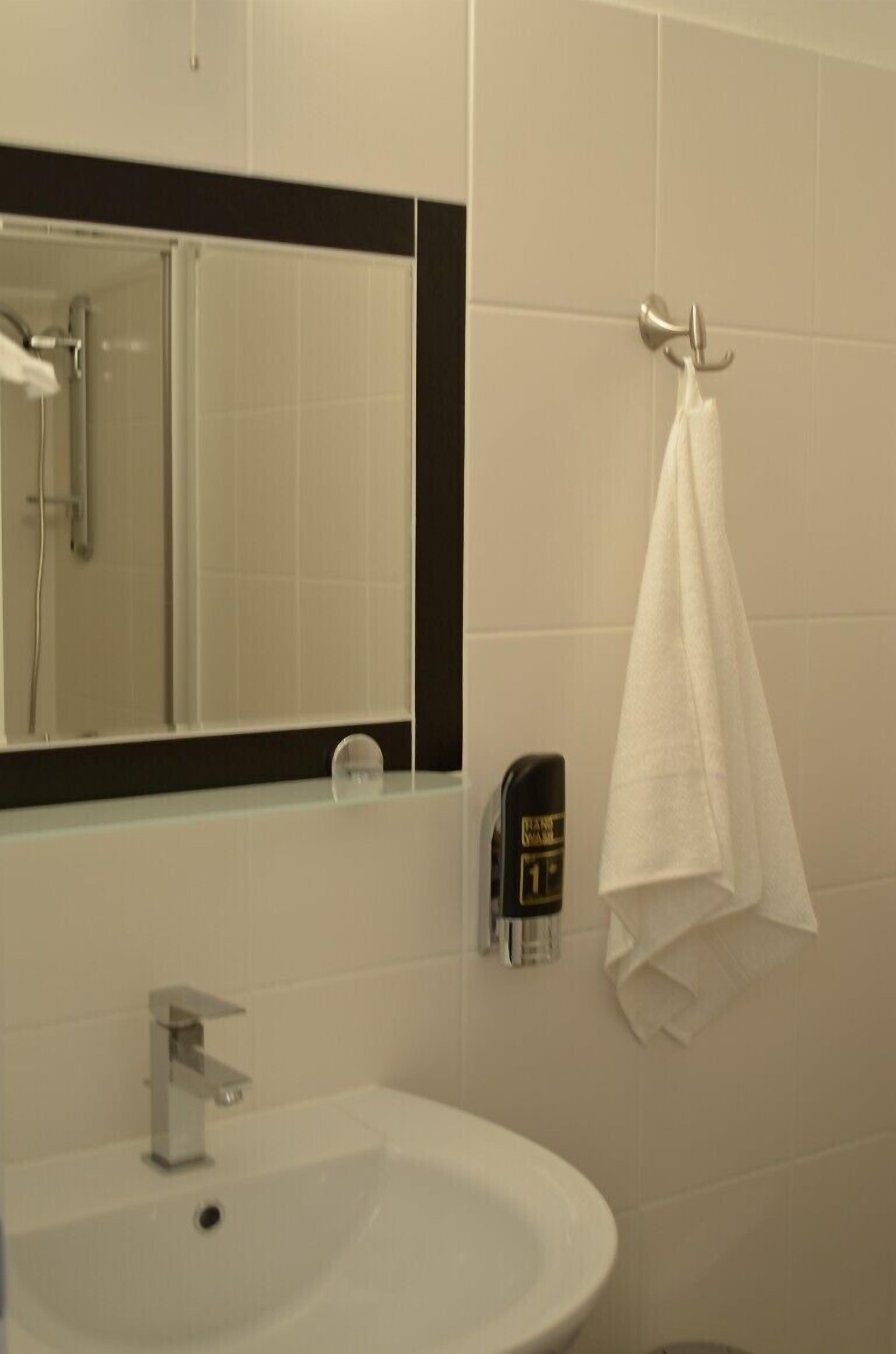 A bathroom with a white sink, a mirror, a towel hanging on a hook, and a soap dispenser on the wall.