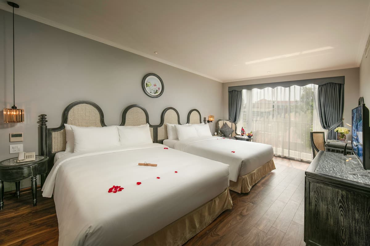 A hotel room with two beds, a wooden floor, a large window with curtains, a small table with a vase, and a wall clock.