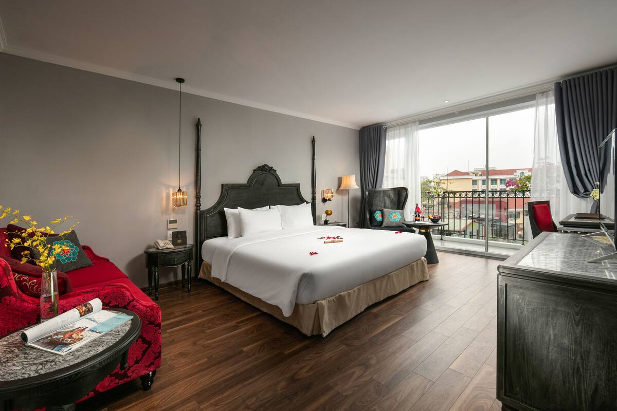 A hotel room with a large bed, a red armchair, a small table with a magazine, a lamp, and a view of the outside balcony.