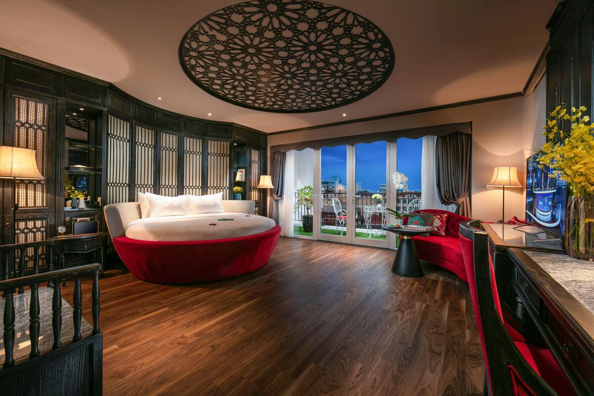 A modern hotel room with a large bed, a red sofa, and a view of the cityscape through large windows.