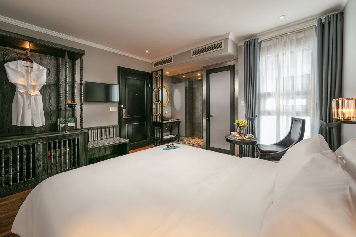 A hotel room with a large bed, a seating area with a chair and a small table, a television mounted on the wall, and a large window with curtains.