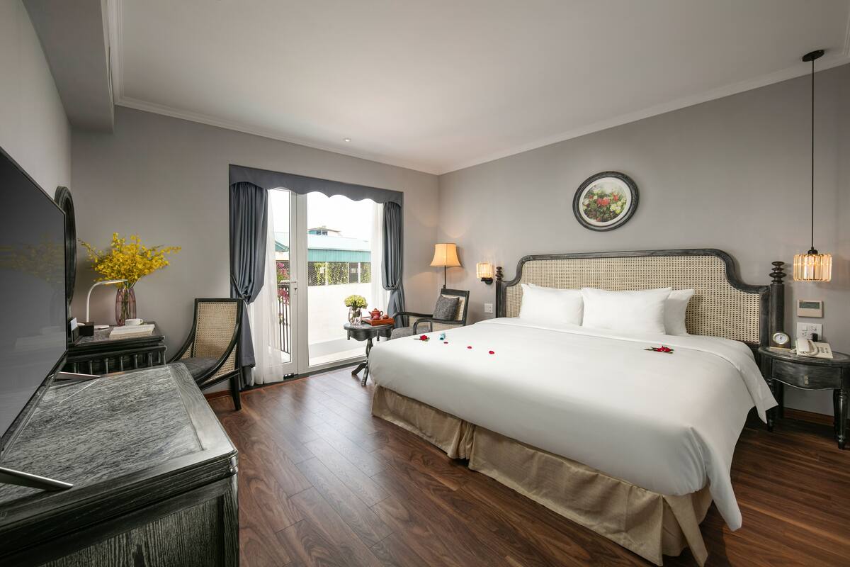 A hotel room with a large bed, a desk with a lamp, a chair, a small table with a vase of flowers, and a view of the outdoors through a window.