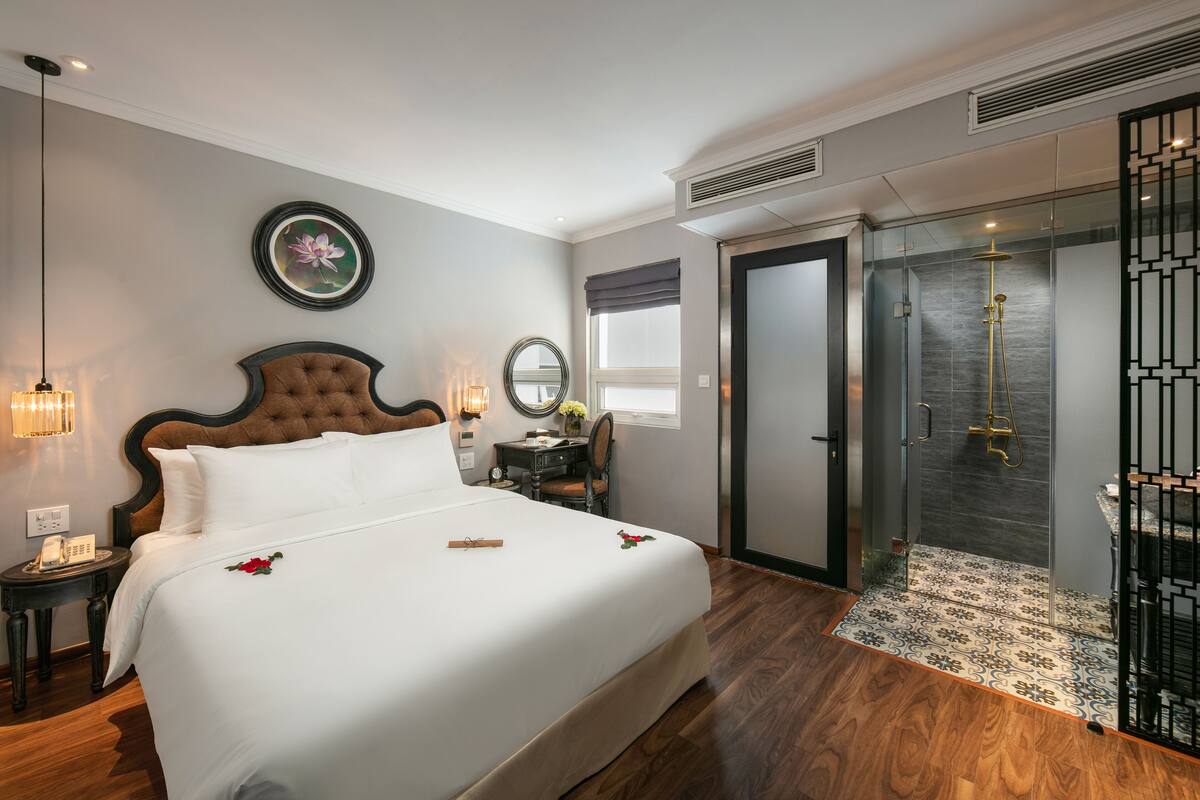 A hotel room with a large bed, a bedside table with a lamp, a mirror, a window with a view, and a shower area.