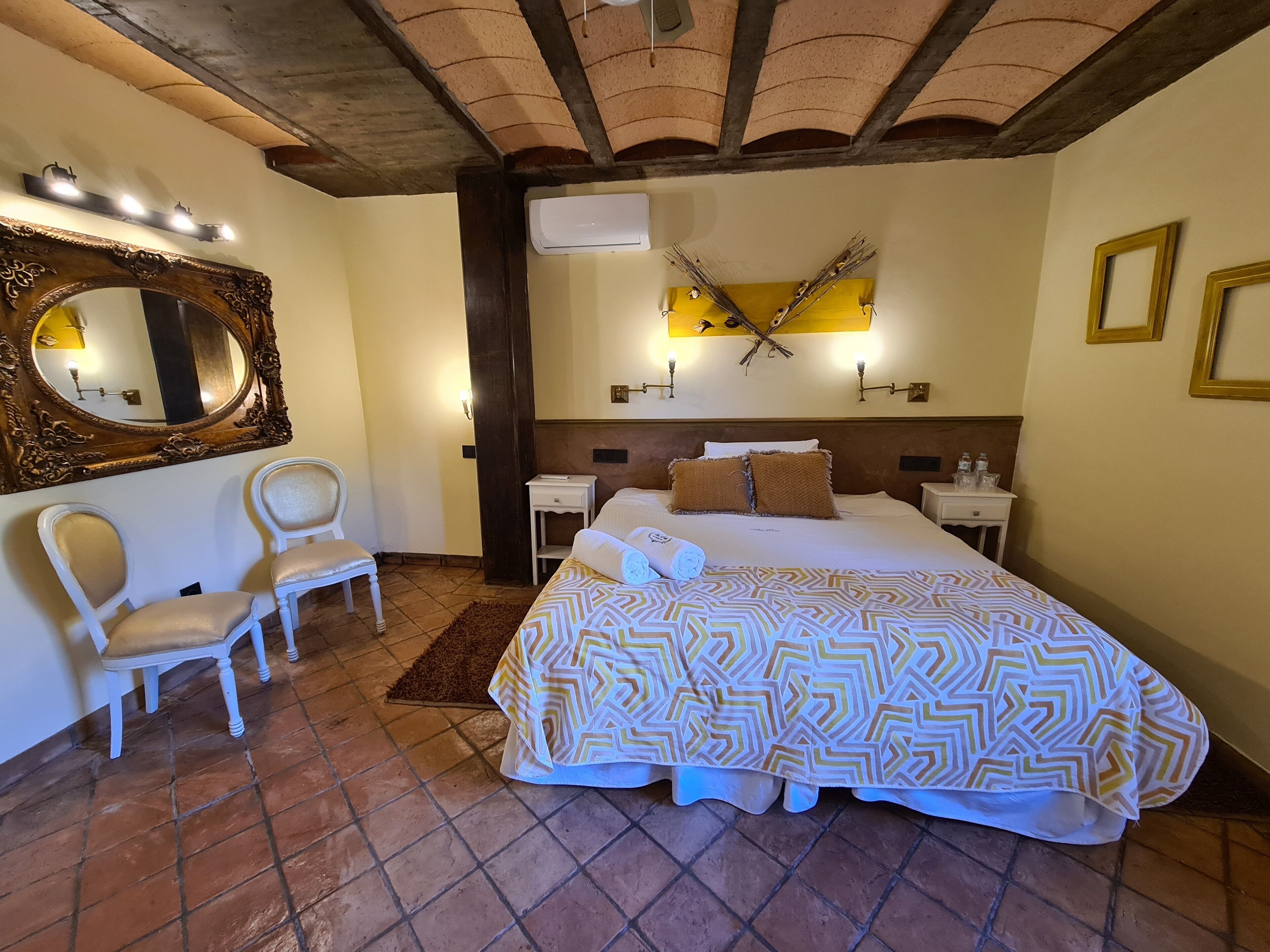 Habitación de hotel con una cama grande, dos sillas, un espejo, una mesita de noche y un aire acondicionado instalado en la pared.