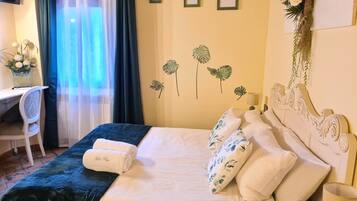 Una cama bien hecha con una manta azul y almohadas blancas, un escritorio con una silla, un televisor montado en la pared y una ventana con cortinas azules.
