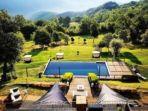 A pool with a wooden deck, surrounded by green grass and trees, with a view of hills in the background. - Hotel Rural & Spa Mas Prat (La Vall de Bianya)