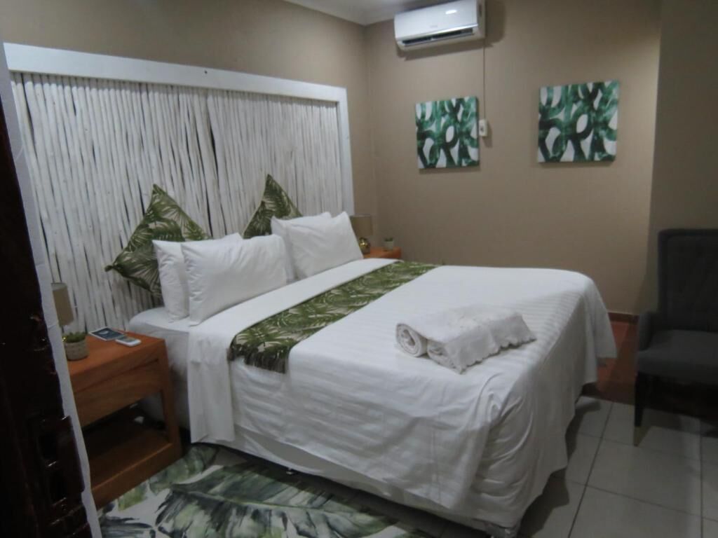 A neatly made bed with white linens and a green leaf-patterned bedspread, a wooden nightstand with a lamp, a wall-mounted air conditioner, and two abstract leaf paintings on the wall.