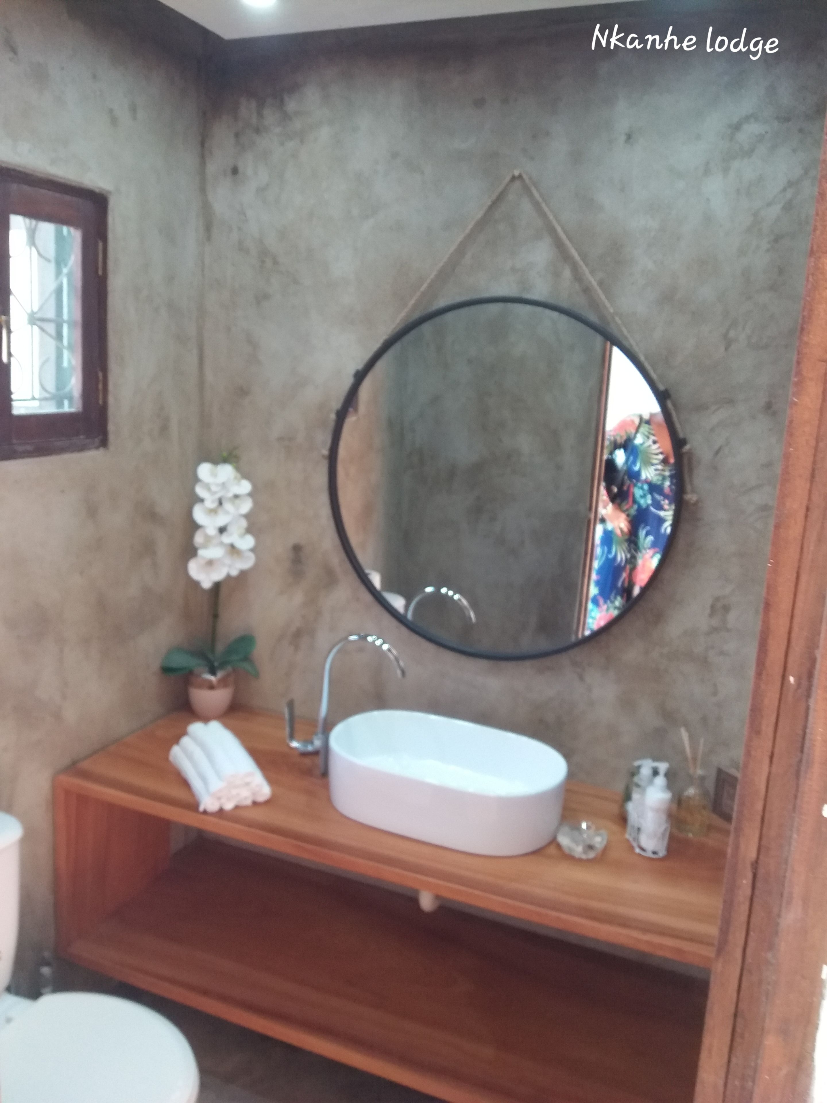 A bathroom with a wooden vanity, a white sink, a round mirror, and a potted orchid.