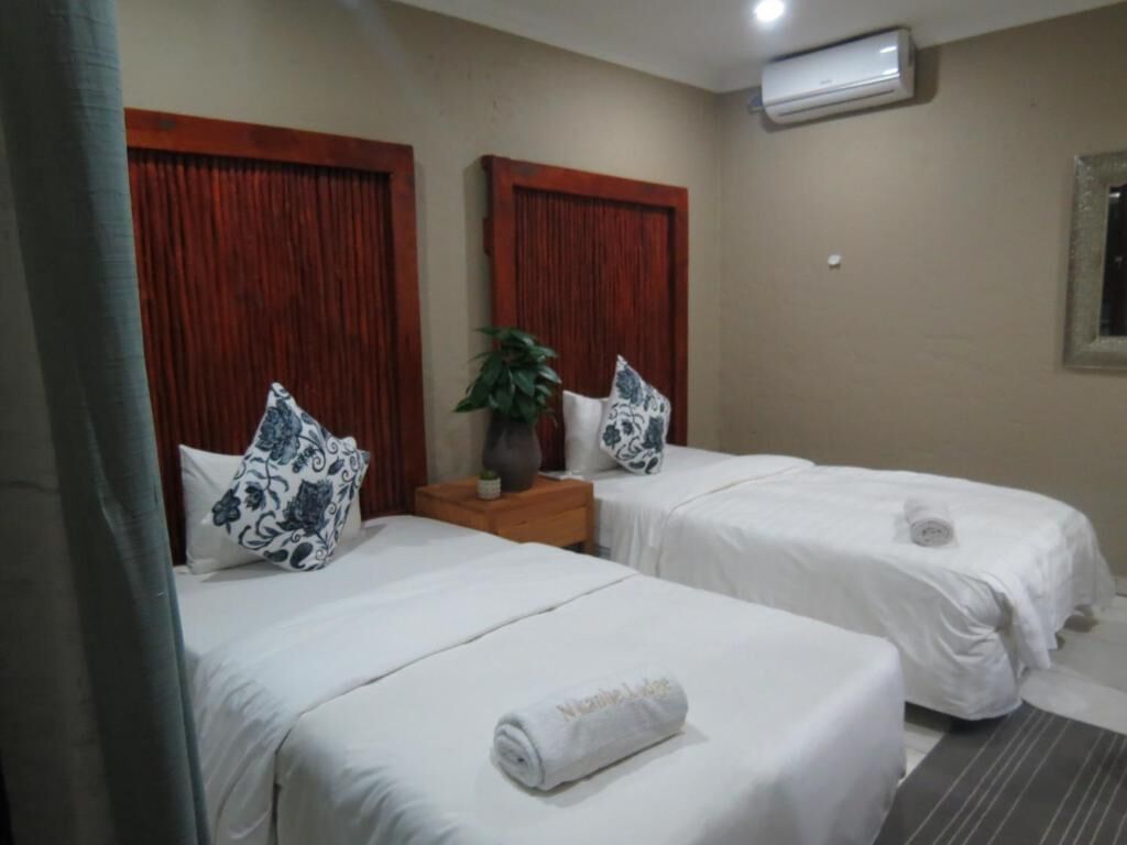 Habitación de hotel con dos camas individuales, cada una con una colcha blanca y una toalla enrollada. Entre las camas, una mesita de madera con un florero y una planta.