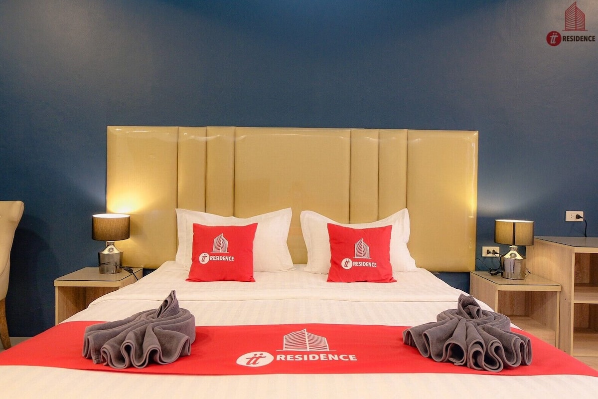 Une chambre d’hôtel avec un grand lit, deux oreillers rouges ornés d’un logo, une serviette rouge posée sur le lit, et deux tables de chevet avec des lampes.