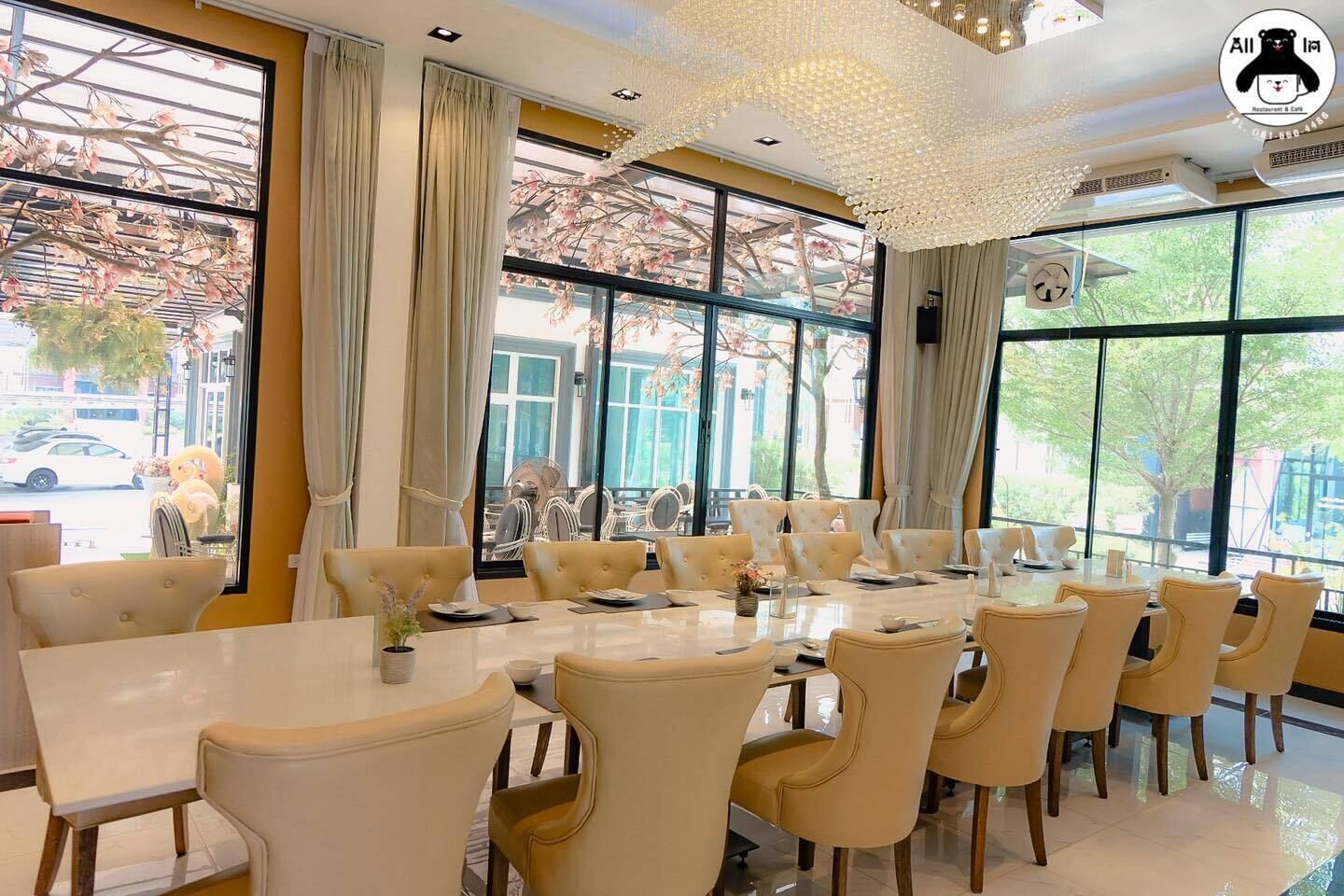 Uma área de refeições com uma mesa comprida e várias cadeiras, janelas amplas com cortinas e um lustre decorativo no teto.