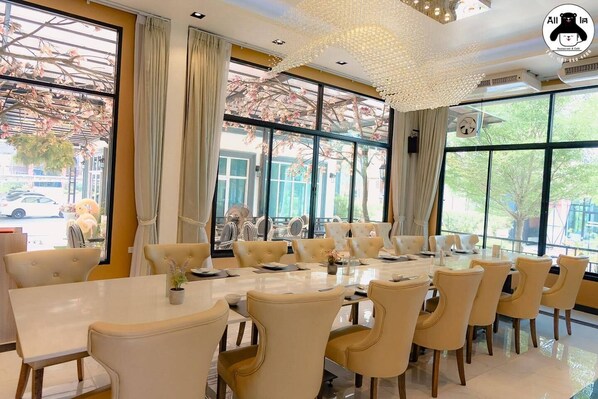 A dining area with a long table and multiple chairs, large windows with curtains, and a decorative ceiling light fixture.