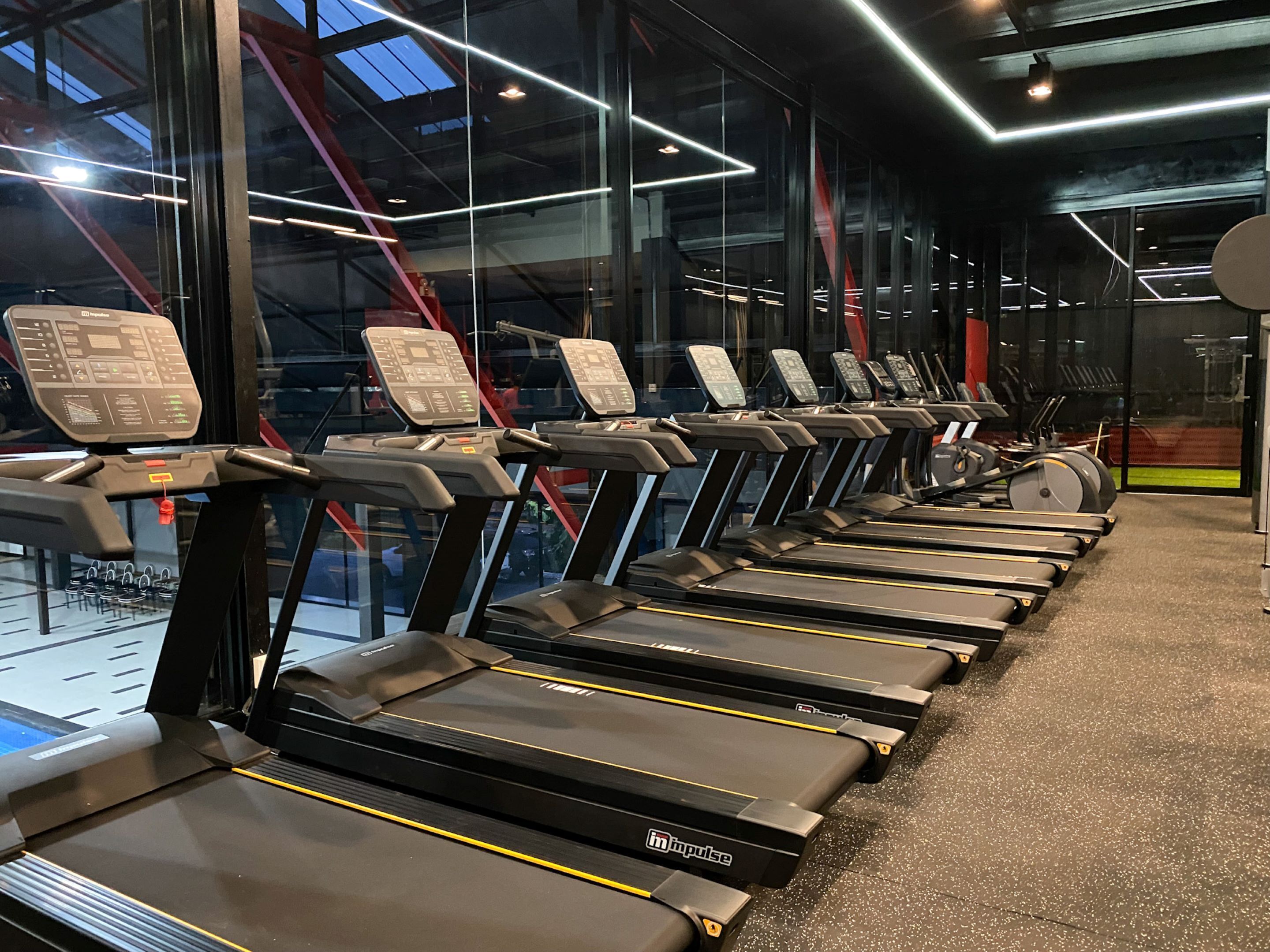 Een rij loopbanden met digitale displays in een sportschool met grote ramen en een hoge plafond.