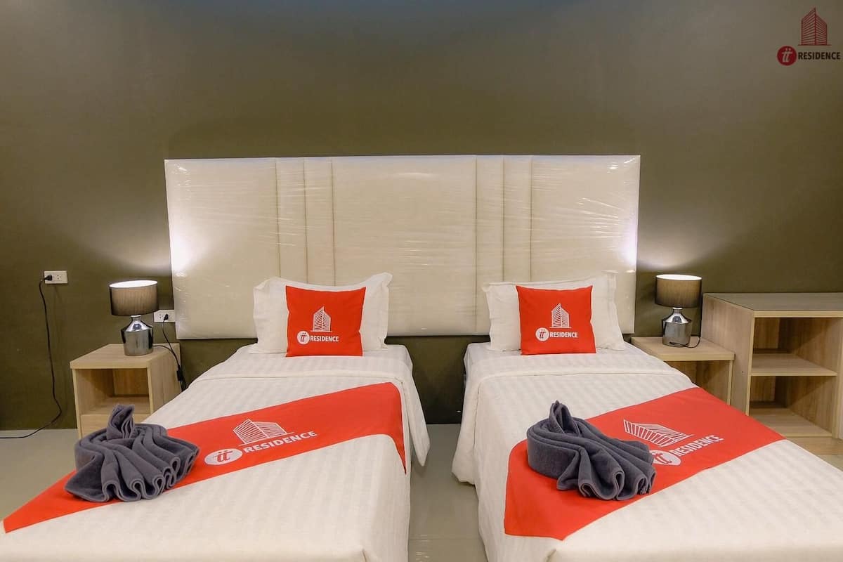 Une chambre d’hôtel avec deux lits, chacun agrémenté d’un oreiller rouge et d’une serviette ornée du logo de l’hôtel. On y trouve des tables de chevet avec des lampes et une petite table en bois.