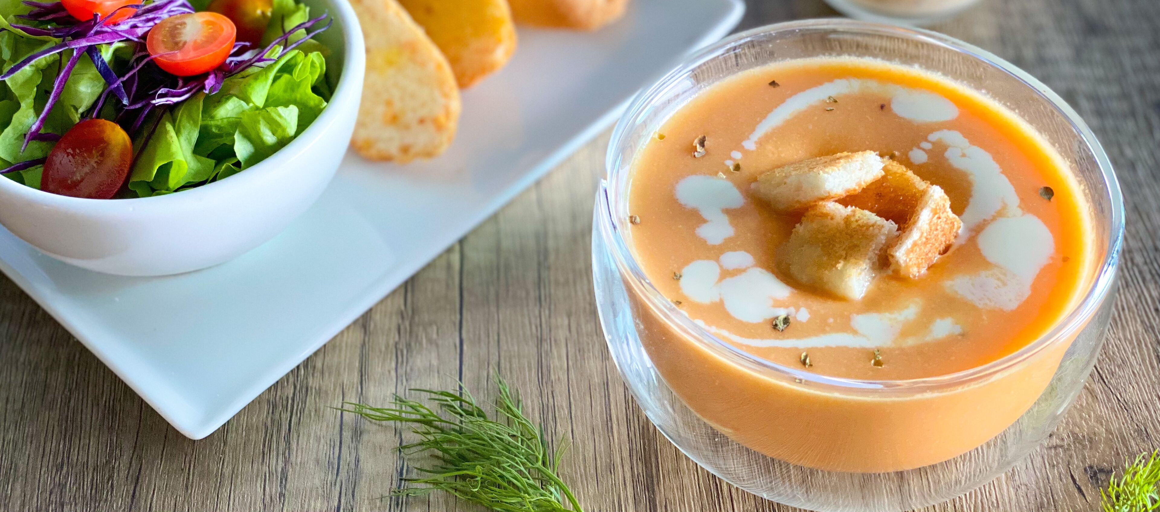 Een kom soep met croutons en een salade op een houten tafel.