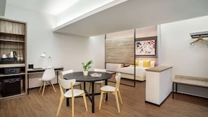 Una sala moderna con una mesa redonda, sillas blancas, un sofá y un banco. Se ve un estante de madera con electrodomésticos y una cafetera. En la pared, hay un cuadro.