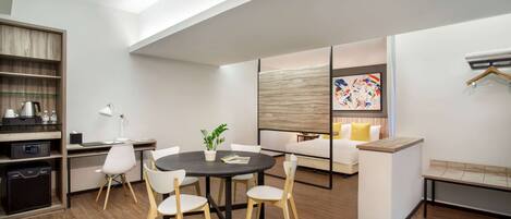 A modern living room with a round table, white chairs, a sofa, and a bench. There is a wooden shelf with kitchen appliances and a coffee maker. A painting is visible on the wall.