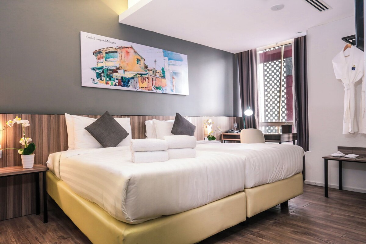 A modern hotel room with a large bed, a desk, a chair, a robe, and a painting of Kuala Lumpur.