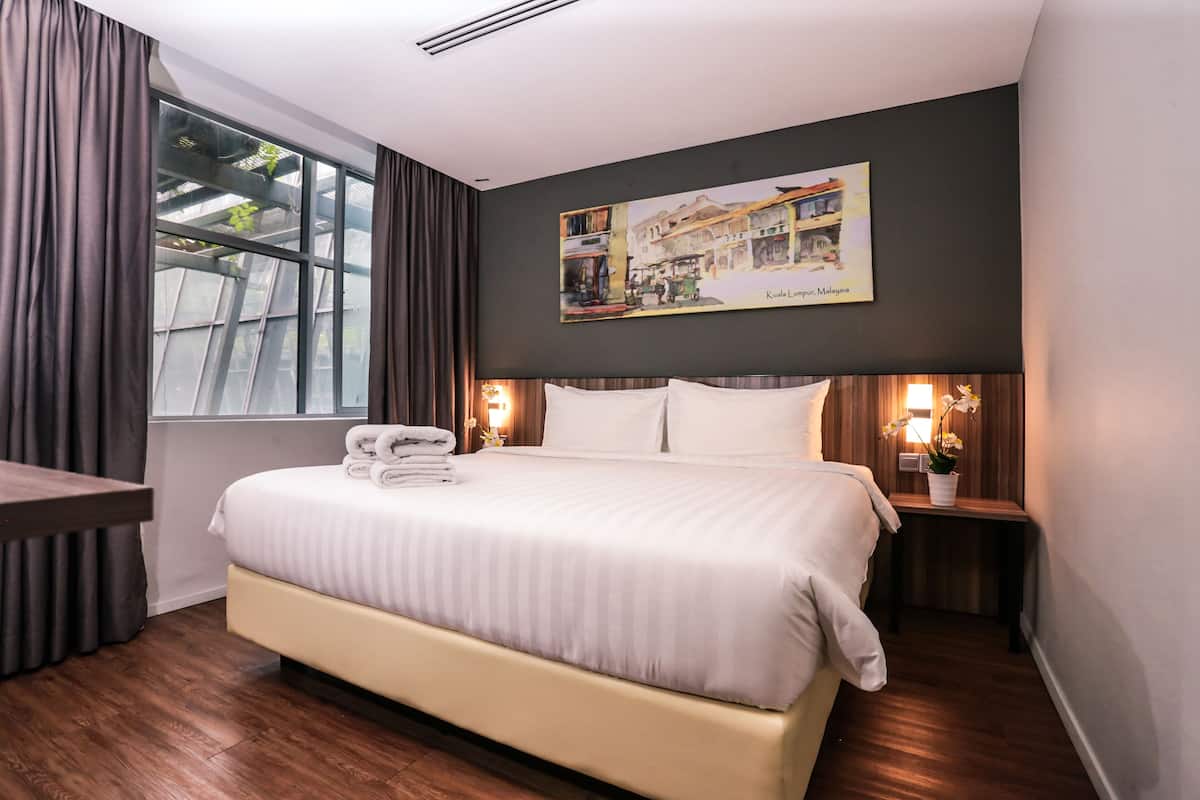 Una habitación de hotel con una cama grande, una mesita de noche, una lámpara y cortinas en la ventana, además de un cuadro colgado en la pared.