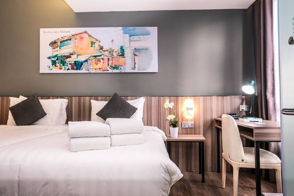 Una habitación de hotel con una cama grande, dos almohadas, un escritorio con una silla, una mesita de noche con una lámpara y un cuadro en la pared.