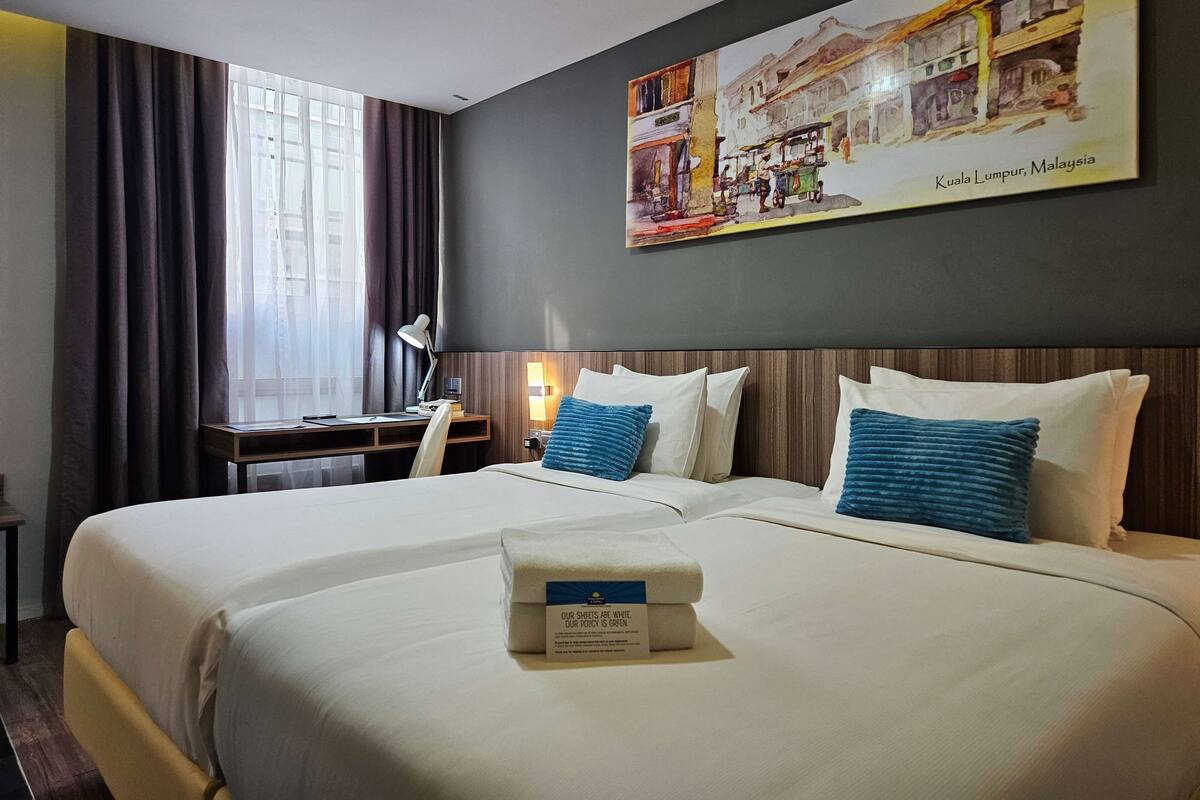 Habitación de hotel con una cama grande, dos almohadas, una mesita de noche con lámpara, un escritorio y una ventana con cortinas.
