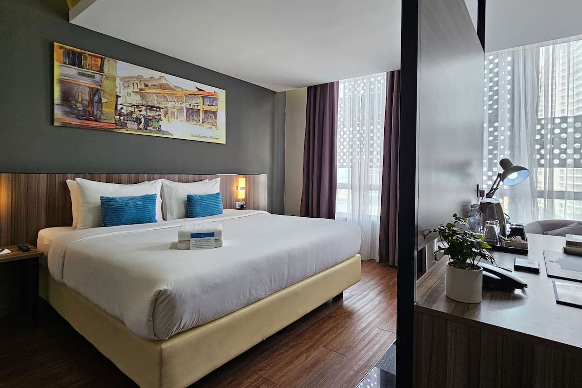 Una habitación de hotel con una cama grande, un escritorio con lámpara, una planta en maceta y un cuadro en la pared.