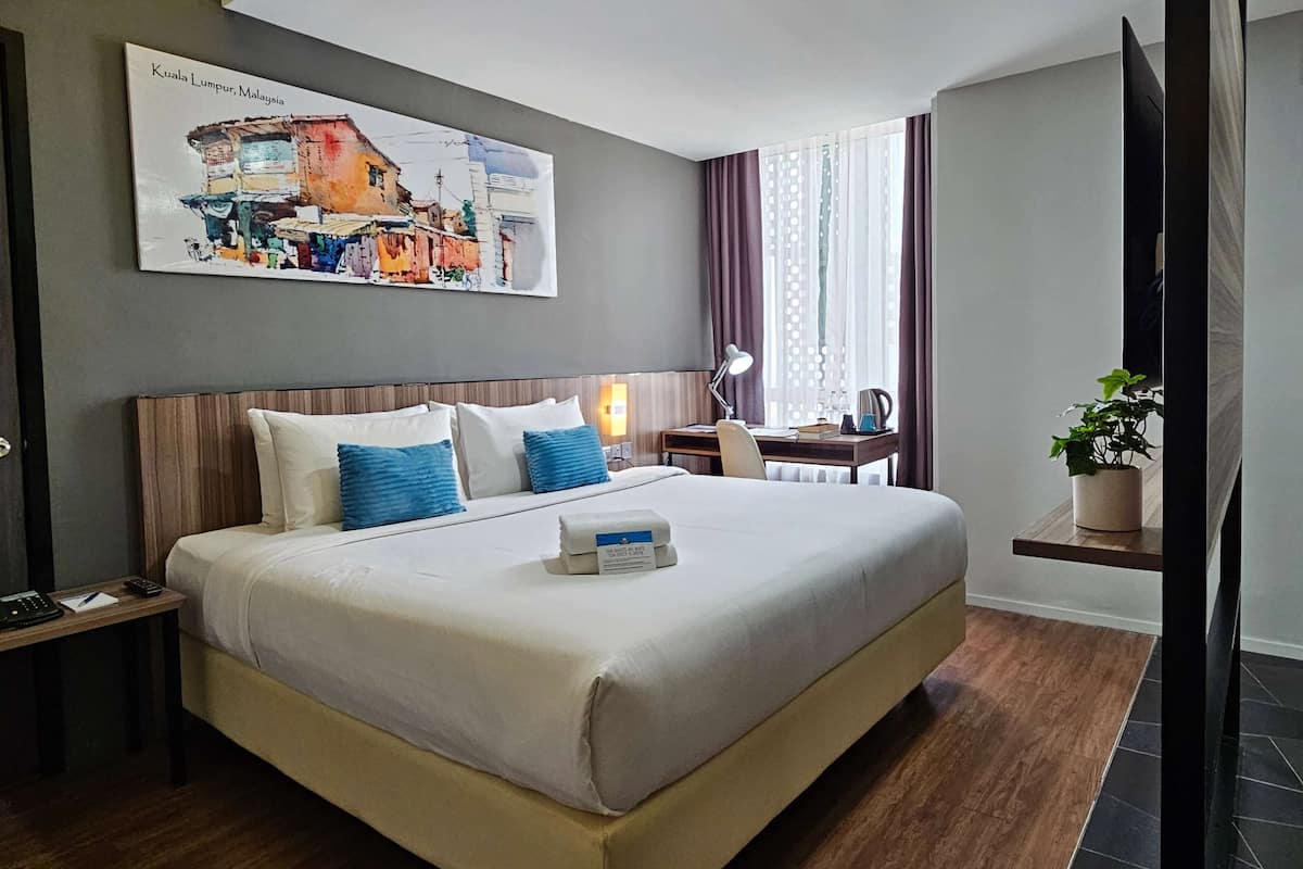 Habitación de hotel con una cama grande, un escritorio con lámpara, una planta en maceta y un cuadro de Kuala Lumpur en la pared.
