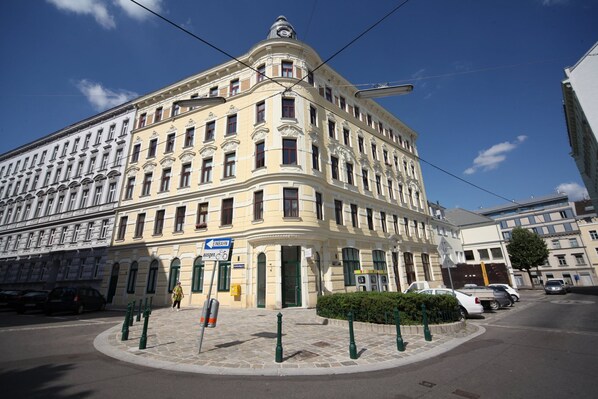 Un bâtiment historique avec une entrée située à l’angle, un panneau indiquant un arrêt de bus et un trottoir pavé.
