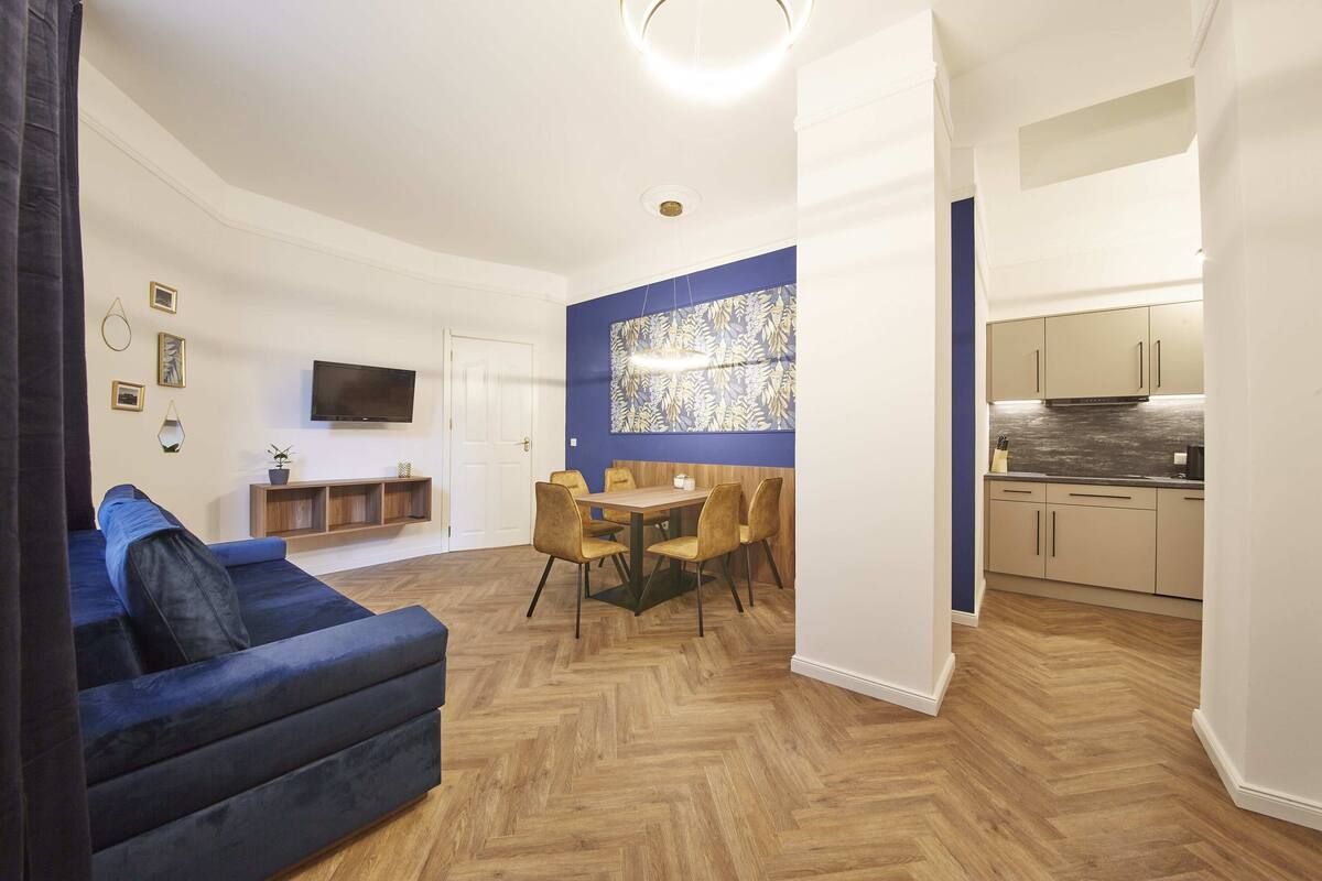 A modern living room with a blue sofa, a dining area with a wooden table and chairs, a kitchen with light-colored cabinets, and a large TV mounted on the wall.