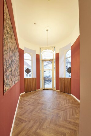Un hall spacieux doté d’un parquet en bois disposé en point de Hongrie, de grandes fenêtres et d’un lustre.